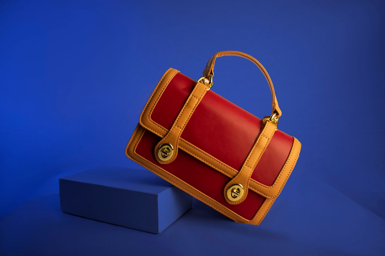 services-02 Stylish red and tan leather handbag with handle and gold accents on vibrant blue backdrop.