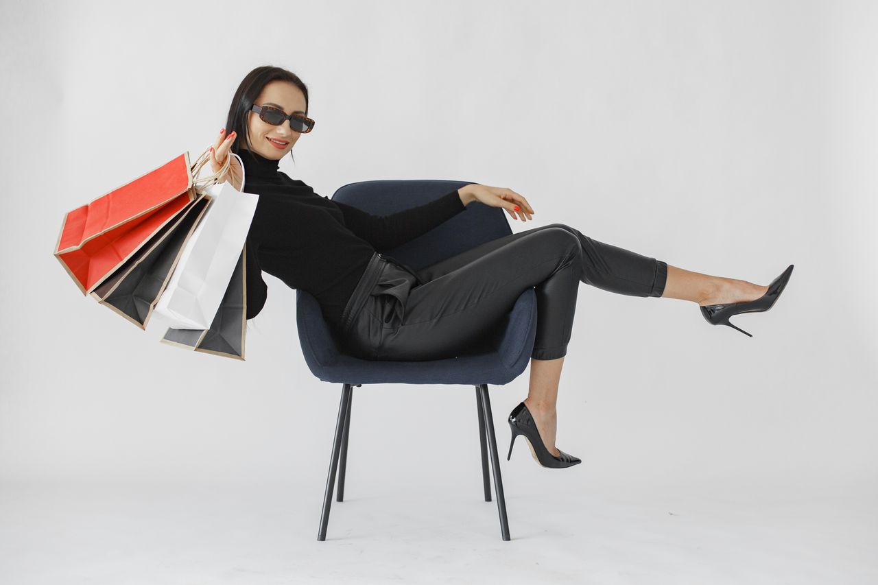 Fashionable woman lounging with shopping bags, wearing stylish sunglasses in a studio setting.